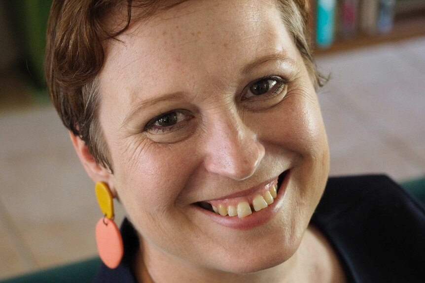 A close-up of a woman with short mahogany hair, round, pastel earrings and a wide smile. Behind her sits a blurred bookshelf.