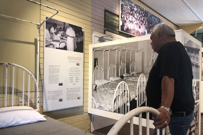 An aging aboriginal man stands in an old dormitory transformed into a museum and reads the captions of historic photographs