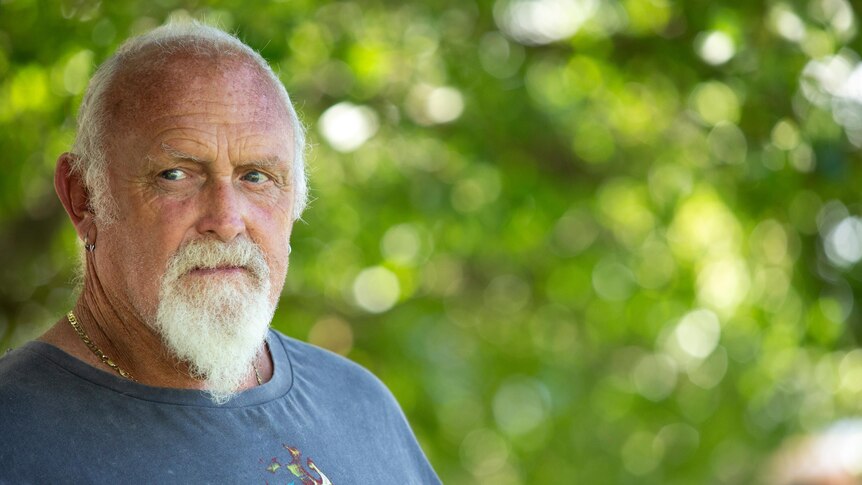 A serious balding older man with grey hair and beard, waers blue tee, looks away from camera. Blurred greenery behind.