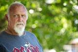 A serious balding older man with grey hair and beard, waers blue tee, looks away from camera. Blurred greenery behind.