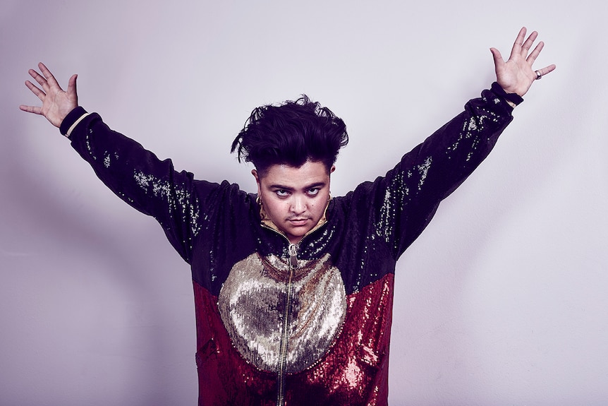 Photo of artist Mojo Juju posing with arms in the air and wearing a sequined jacket featuring the Indigenous flag.