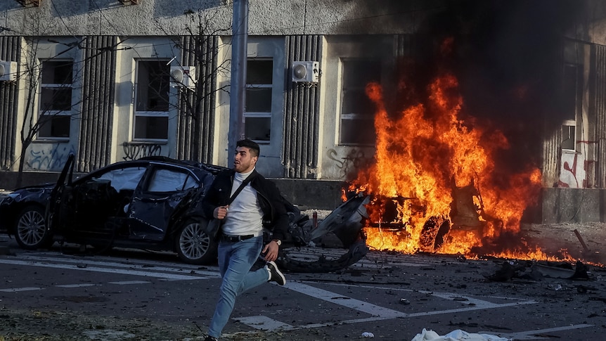 A young man wearing jeans and carring a bag runs past a flaming car in a city street.
