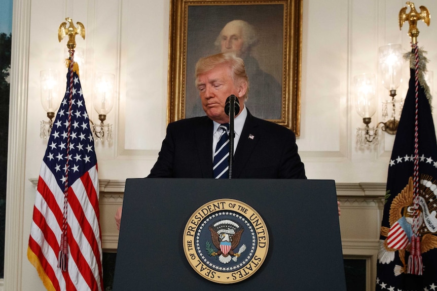 Donald Trump looks downcast as he stands at a podium flanked by flags.