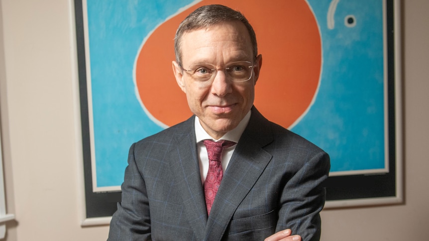 A man with short hair wearing thin framed glasses smiles with arms folded wearing a dark grey suit and maroon tie