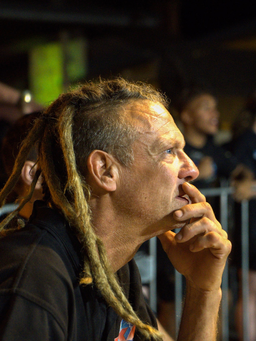 A man with dreadlocks watches a fight with his hand on his mouth.
