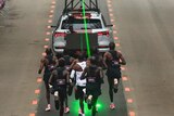 A group of male runners are following a timing vehicle which is projecting a green laser on the ground to guide them