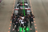 A group of male runners are following a timing vehicle which is projecting a green laser on the ground to guide them