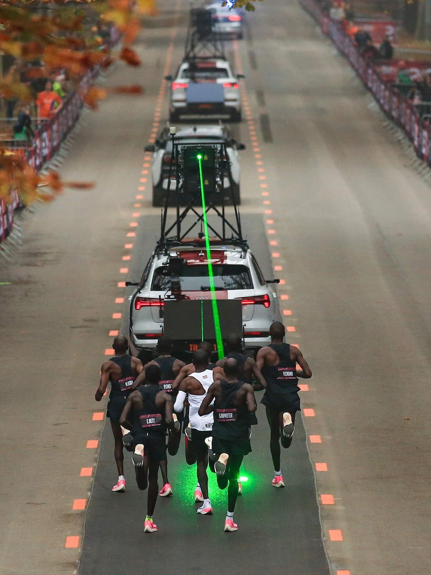 A group of male runners are following a timing vehicle which is projecting a green laser on the ground to guide them