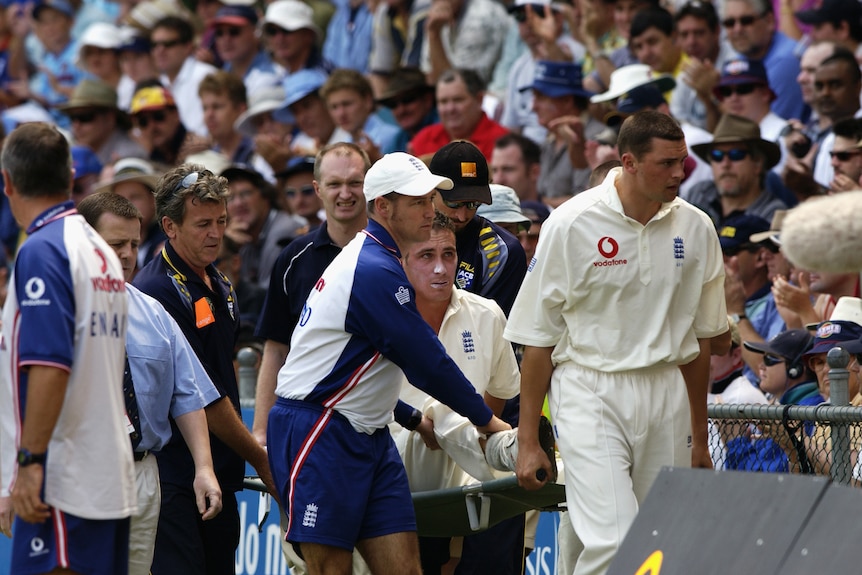 An England cricketer is carried of the ground on a stretcher as a medical assistant holds his injured leg.
