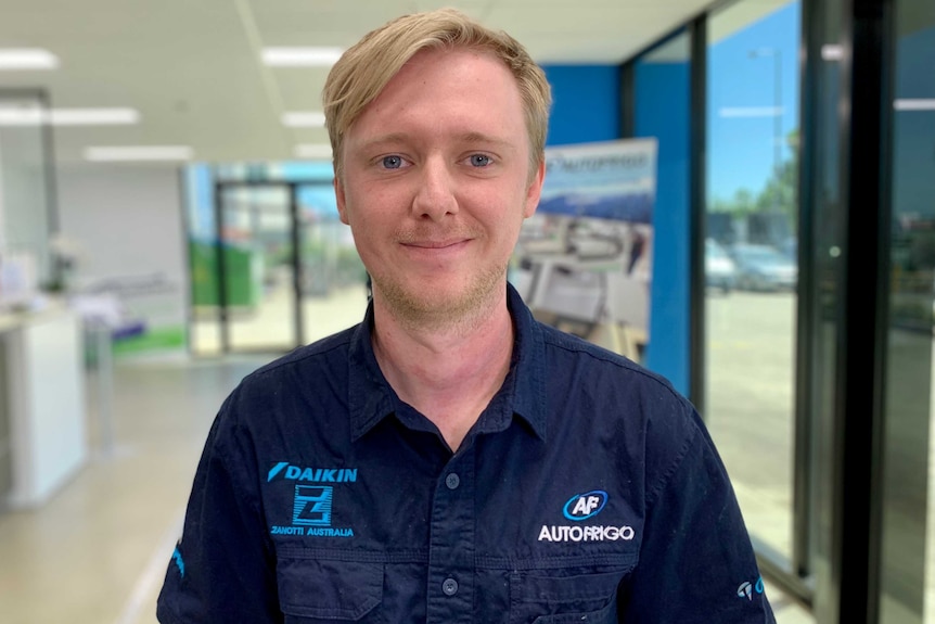 A 29 year old man with blonde hair wearing a dark blue work shirt with 'AutoFrigo' on it, smiles to the camera.