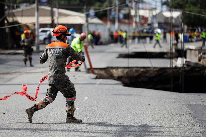 A soldier wearing an orange helmet is running on a road in front of asinkhole with emergency tape. 