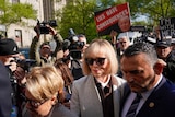 An older blonde woman walks through a crowd of people, one holds a sign with Trump's face and LIES HAVE CONSEQUENCES