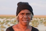 An Indigenous woman in a black top and black beanie looks at the camera, in the outback, background blurred.