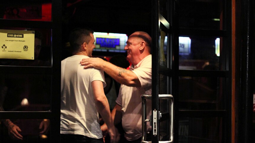 Two middle-aged men embrace each other on the door of a dark pub at night time.