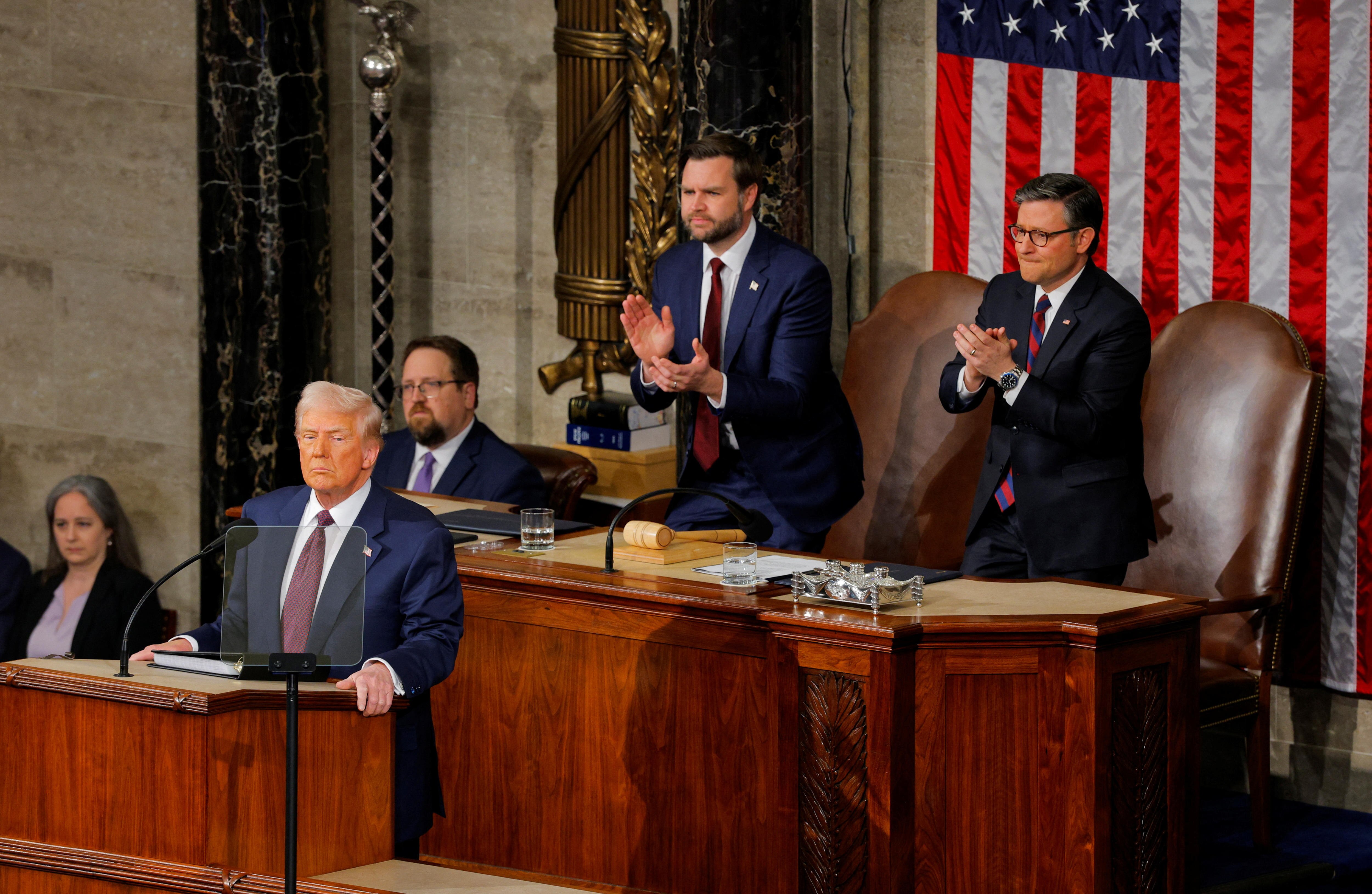 Donald Trump stands at a dais and speaks. Men stand behind him and applaud.