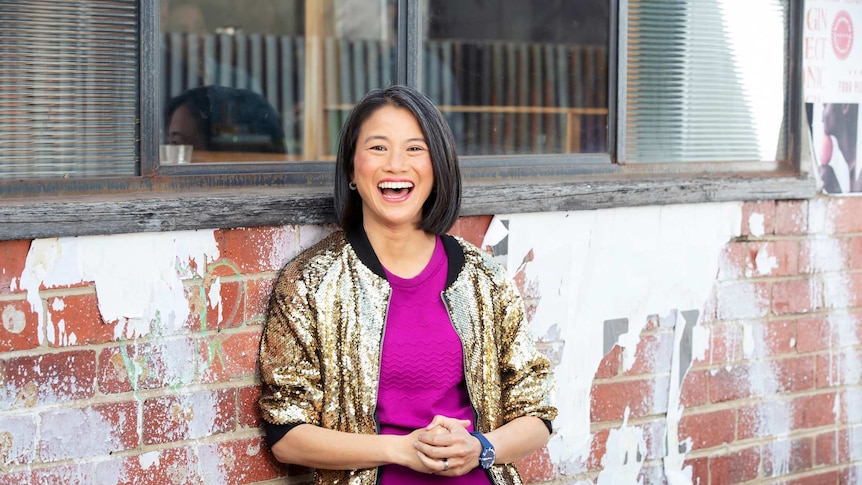 Lisa Leong leaning against a brick wall with windows in it, laughing.