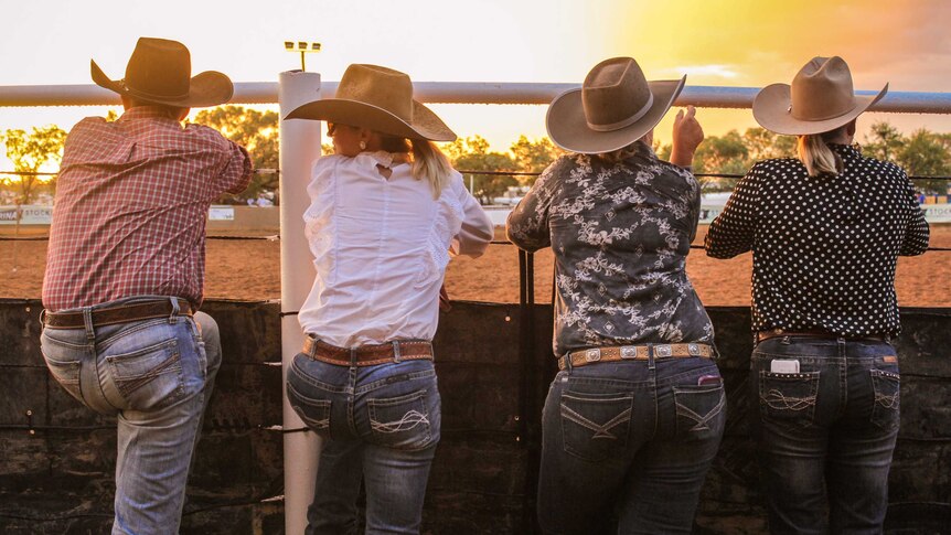 Four people dressed in blue jeans and cowboy hats lean on the arena, with a bright yellow sunset in the background.