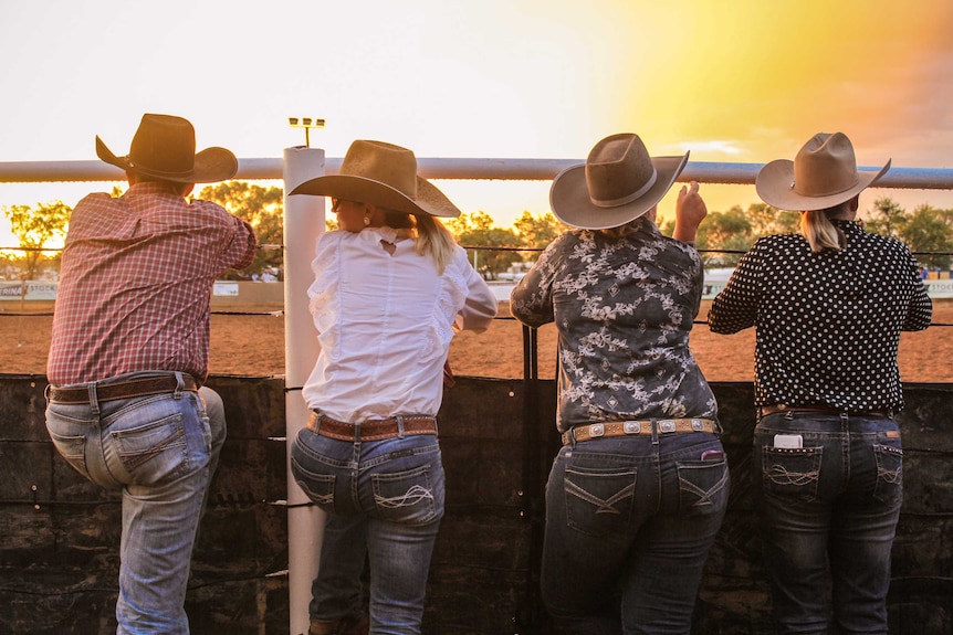 Four people dressed in blue jeans and cowboy hats lean on the arena, with a bright yellow sunset in the background.