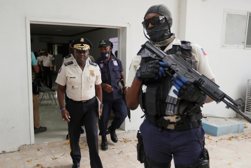 A middle-aged Haitian man in a white police uniform exits a doorway behind a heavily armed guard.