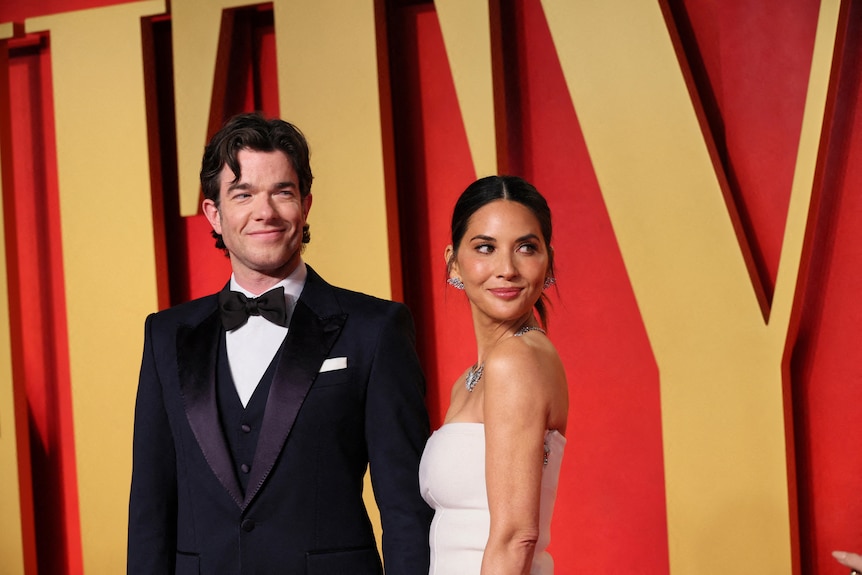 John Mulaney wears a black suit and bow tie, while Olivia Munn wears a strapless white dress