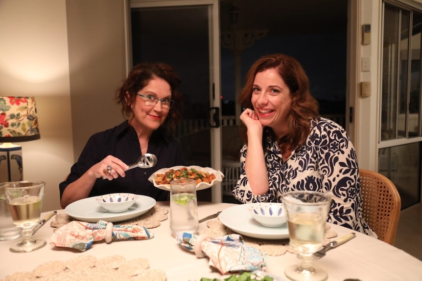 Two women sitting across a dinner table with one pretending to serve up food to the other, both smiling at the camera.