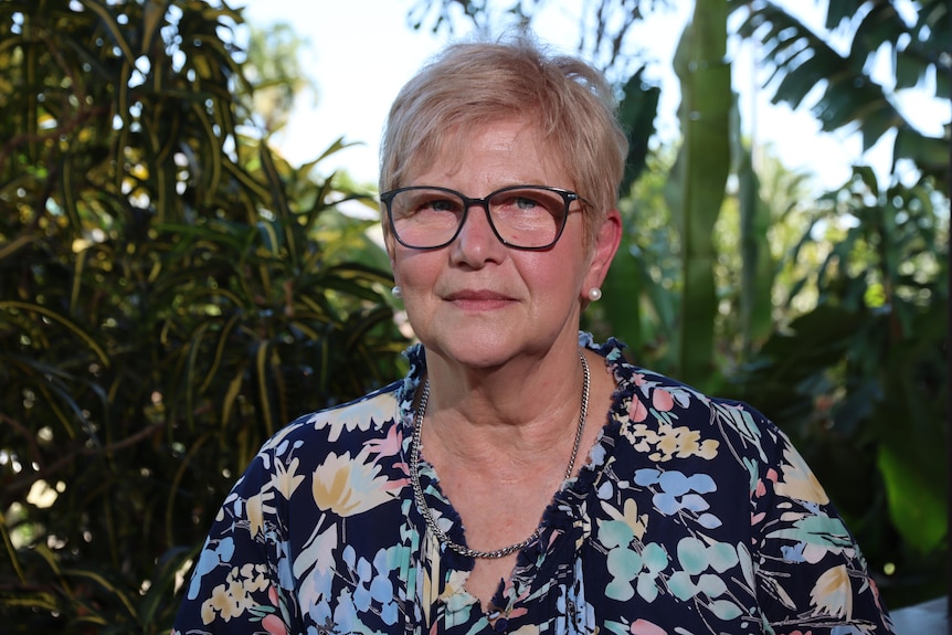 An older woman wearing glasses and a floras shirt stands against a leafy background