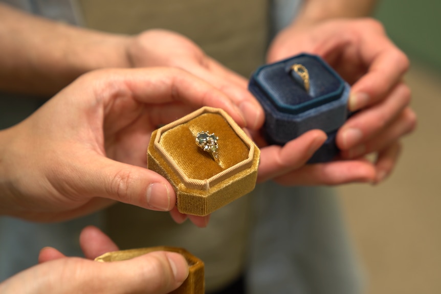 Rohan and Hiep are holding thier finished gold engagement rings. One is in a yellow box and the other in a navy blue box.