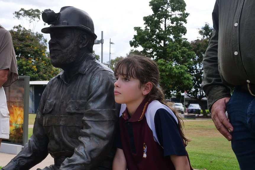 Young girl sits beside statue of a coal miner, he mother stands beside her. 
