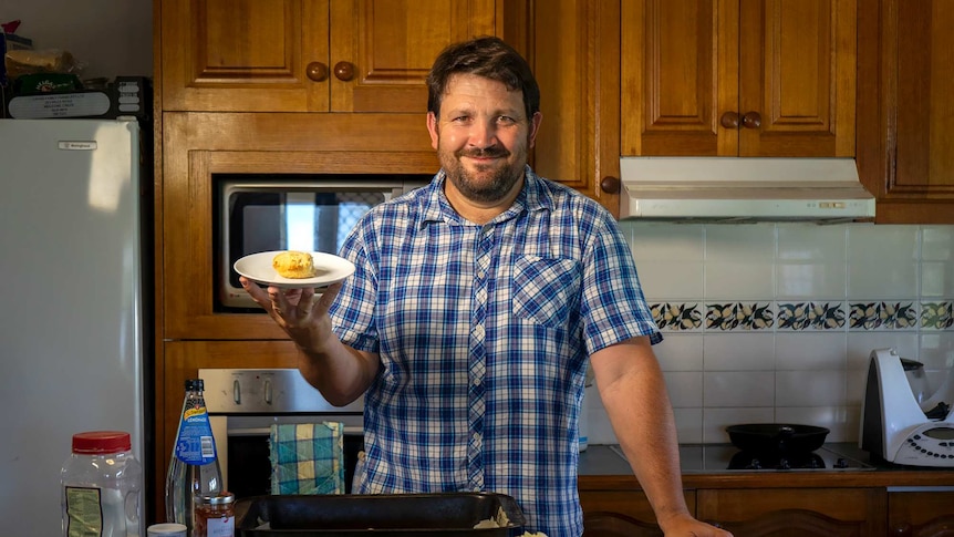Ben Turnbull stands in the kitchen holding a plate with a scone on it; scone-making ingredients on the counter in front of him.
