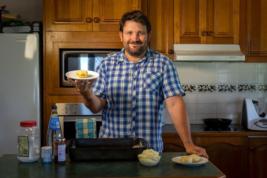 Ben Turnbull stands in the kitchen holding a plate with a scone on it; scone-making ingredients on the counter in front of him.