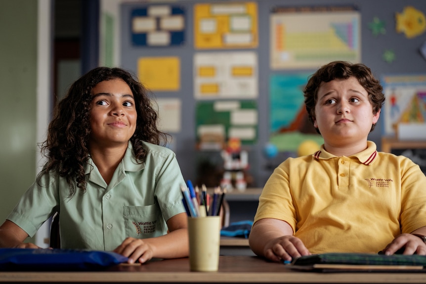 Two boys sit at a desk in a school room, looking up mischeviously.