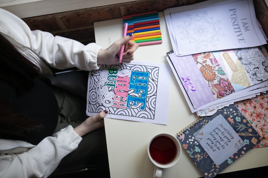 A woman uses pens to colour a colouring-in page with the words 'Life is tough but so are you' on it.