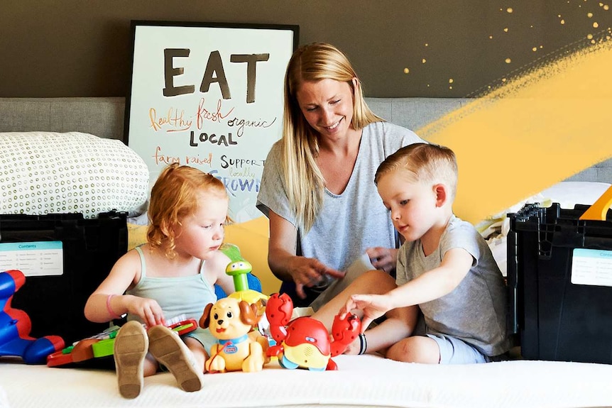 A woman is sitting on a bed with two young children. The kids are playing with toys and they have boxes around.