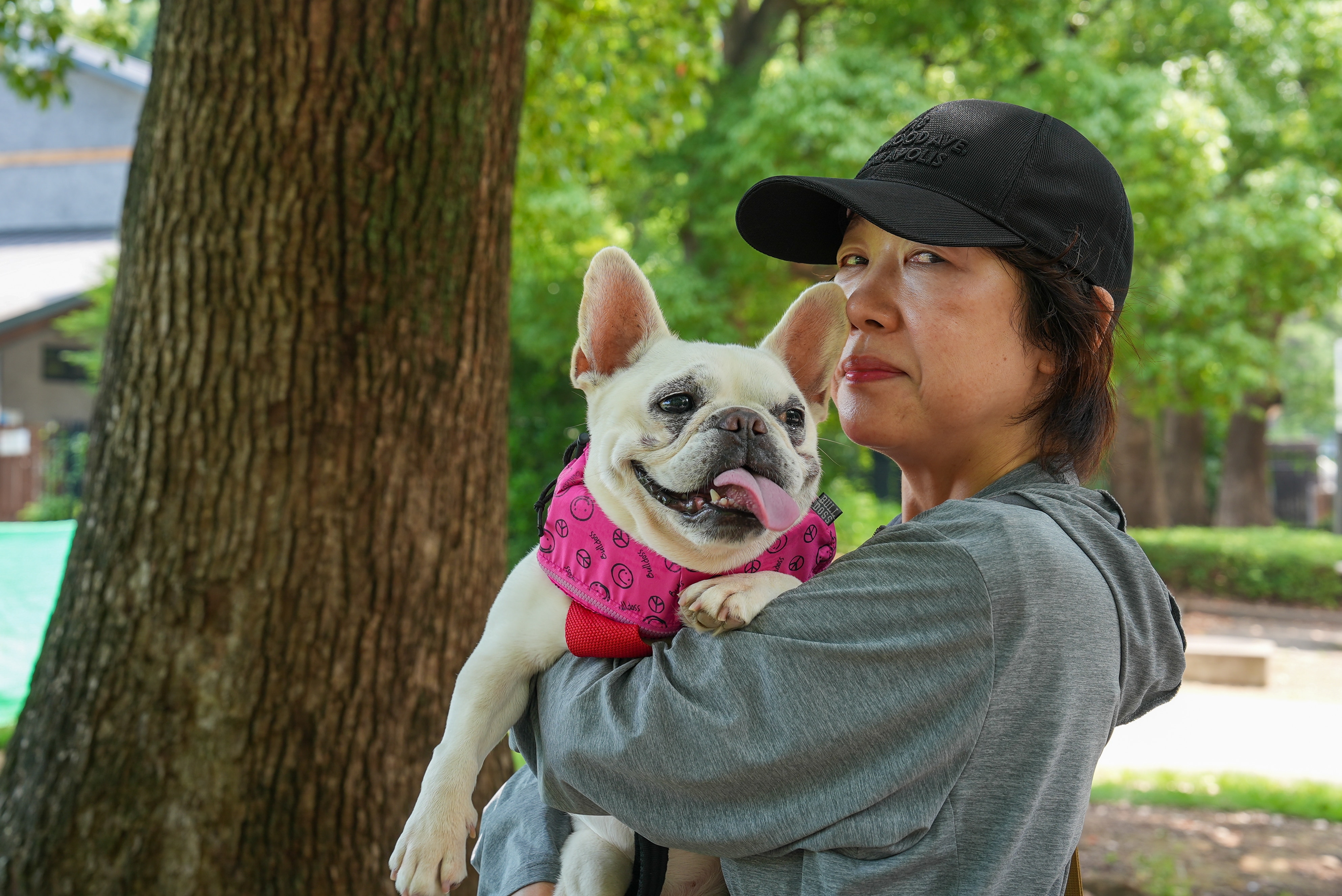 A woman in a grey hoodie and black cap holds a white pug dog in a pink jacket.