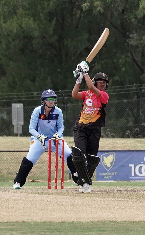A female cricketer hits the ball while a light blue keeper watches.