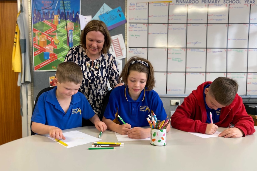 A woman stands behind three children sitting at a desk and colouring in.