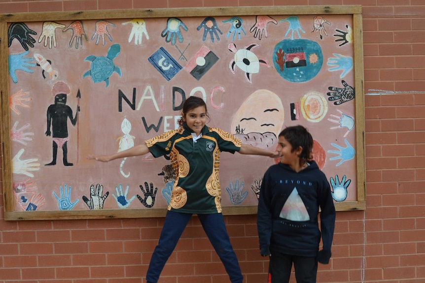 A young student stands in front of a NAIDOC mural.