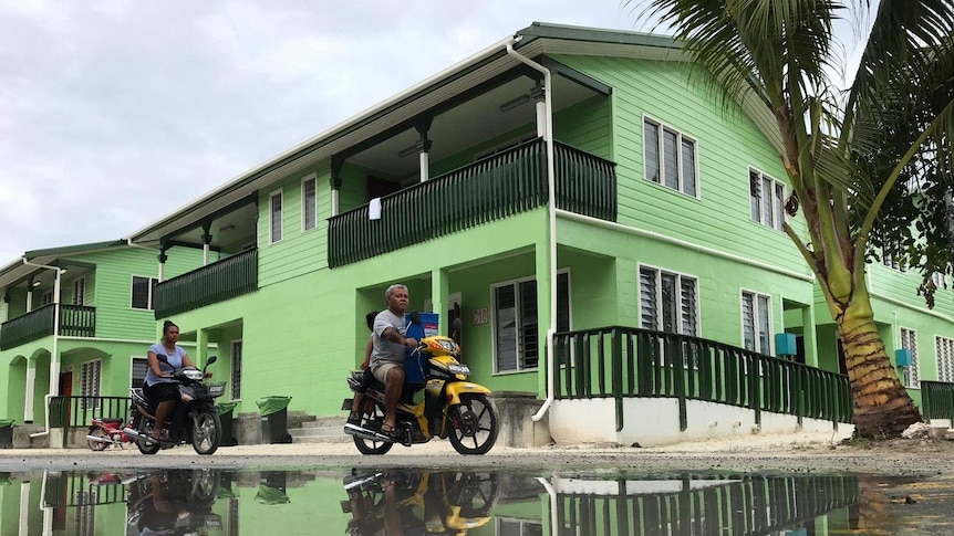 A man and a woman rides a motorbike beside a puddle, in front of a green house.
