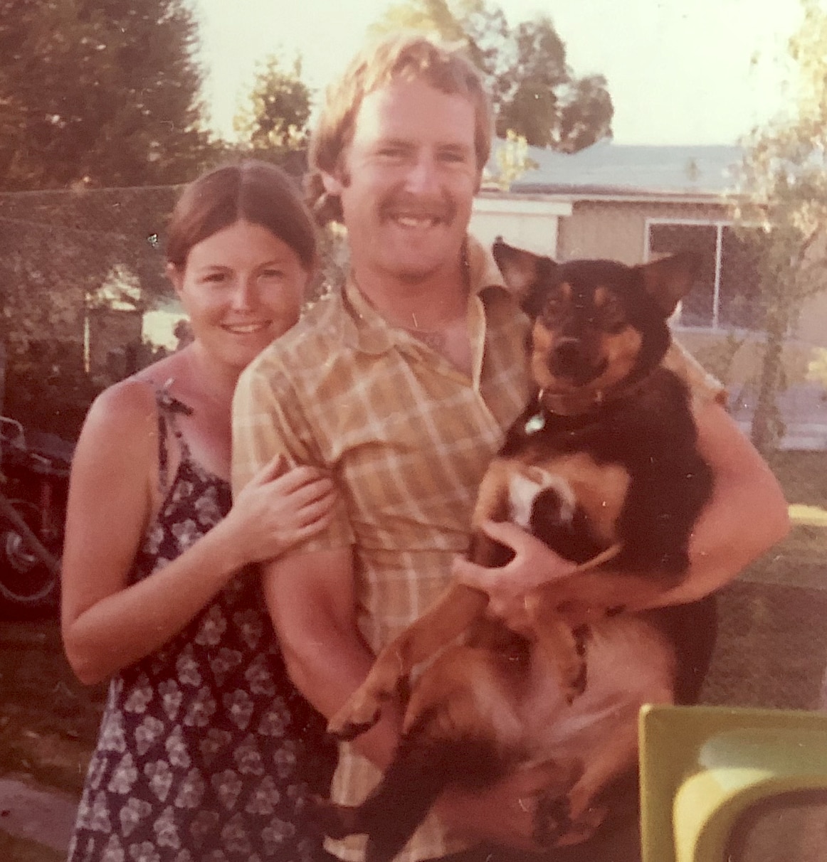 A young woman and man stand in a garden and smile. The man is holding a dog. 