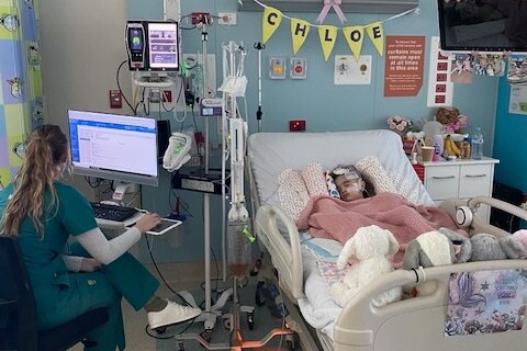 A small girl rests in a hospital bed while a healthcare worker checks a computer nearby, a sign hung over the bed reads "Chloe".