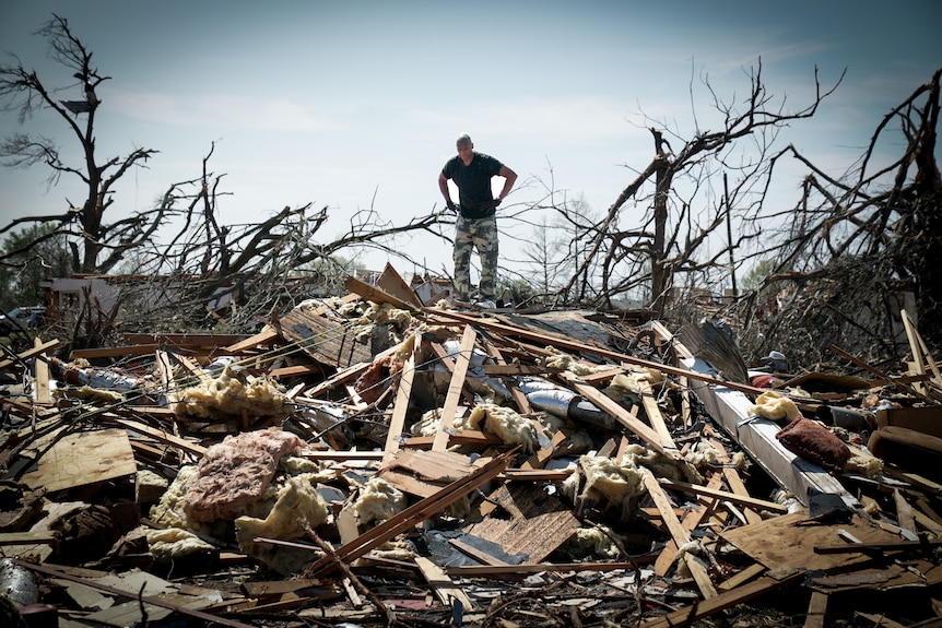 A man in a t-shirt stands in the rubble of his town 