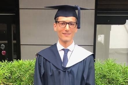 Damion stands in a university gown in front of the entrance to a building.