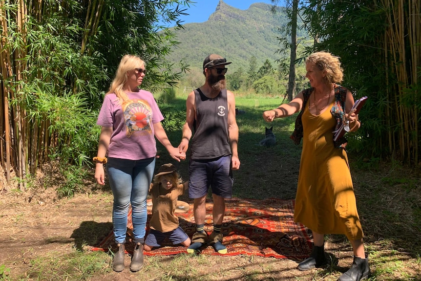 TWo people holding hands with a small child between them, a marriage celebrant off the the right.