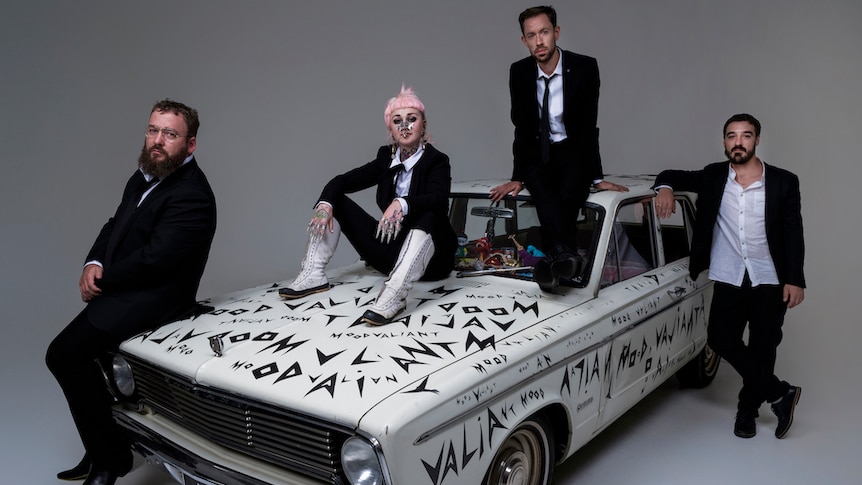 Hiatus Kaiyote wearing suits posing on and around a white Holden Valiant with Mood Valiant written all over it