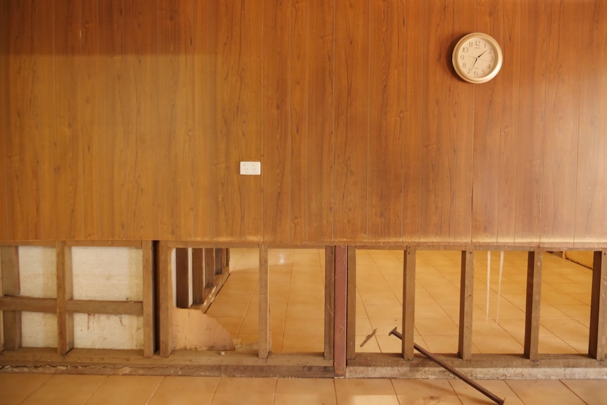 A motel room with a clock on the wall. There is damage to the bottom part of the wall. 