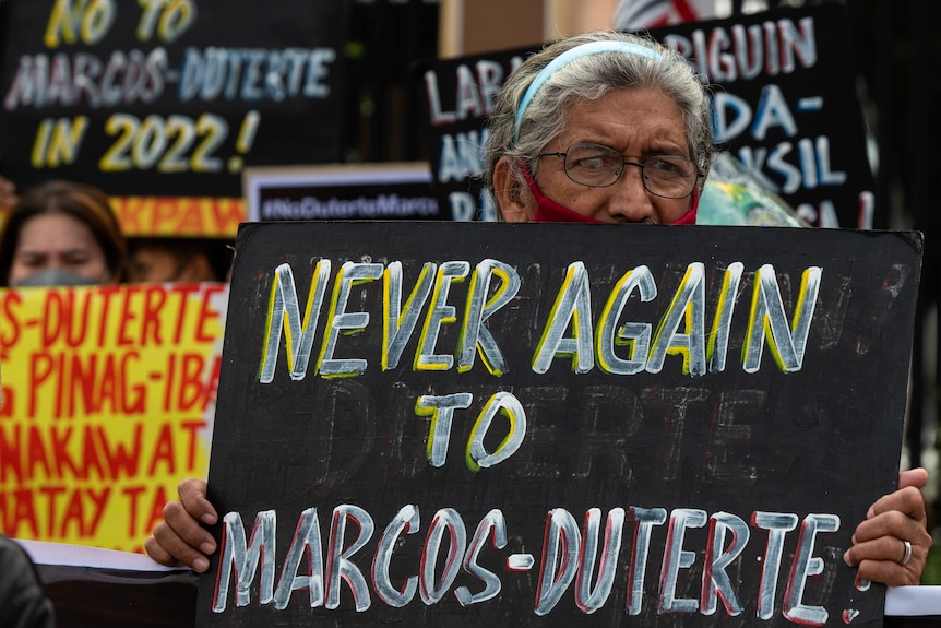 A older woman holds up a placard that says 'never again to Marcos-Duterte'