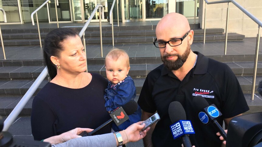 A bald man with glasses and beard outside the Perth District Court, alongside a woman holding a small child.
