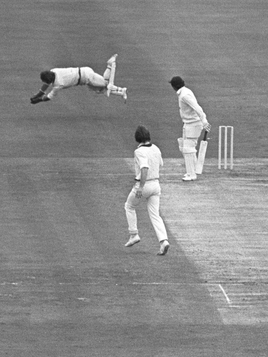 Rod Marsh dives to his right and catches a ball with two hands as batter and bowler look on