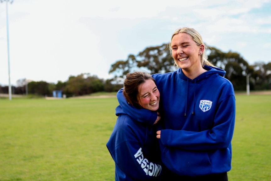 Two young women smile wrestling wearing blue hoodies on a football oval