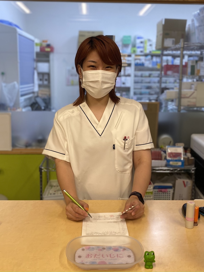 A woman wearing white scrubs and a white face mask sits at a pharmacy counter. 